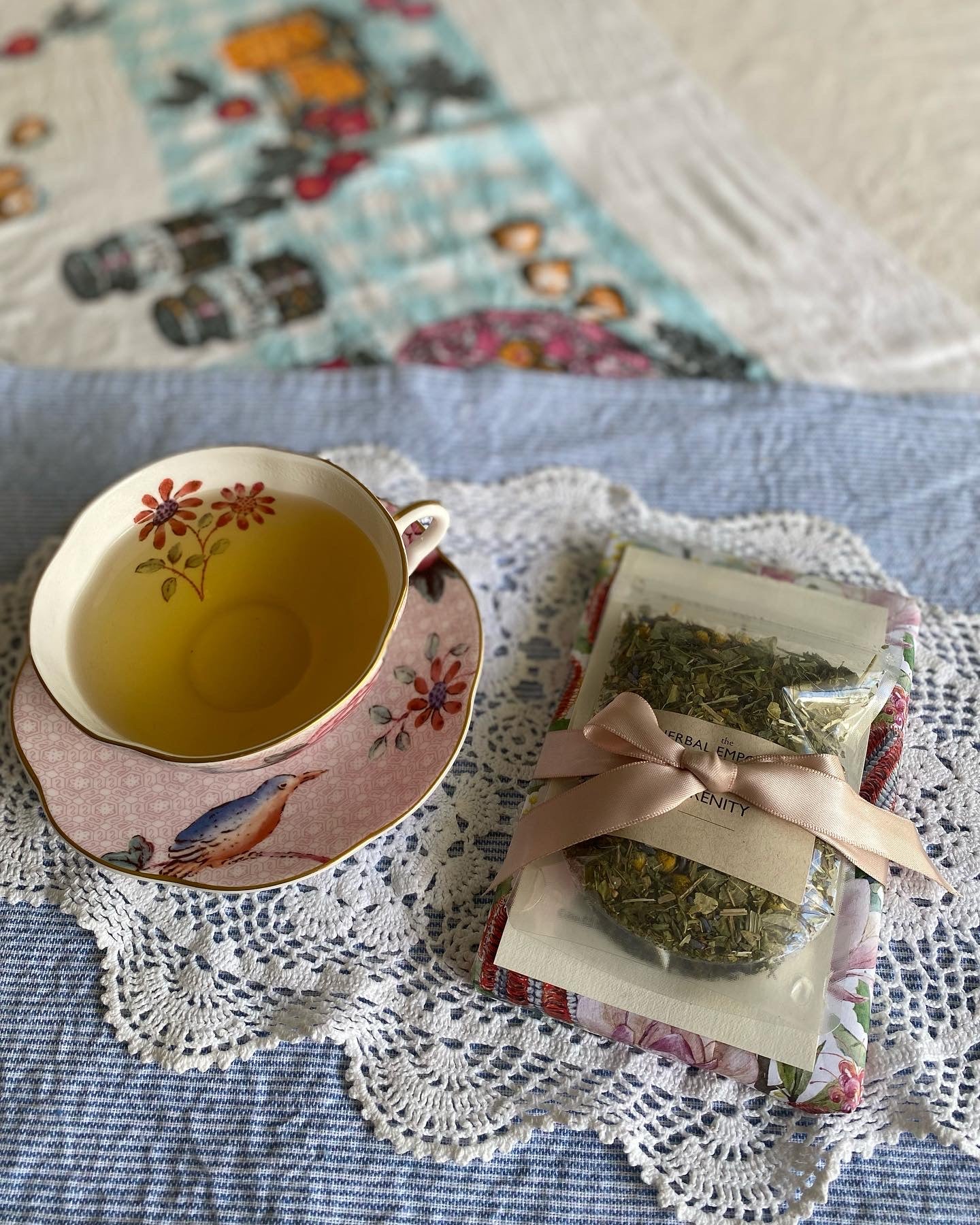 Teacup with floral design on a doily, next to a package of tea leaves on a quilted surface.