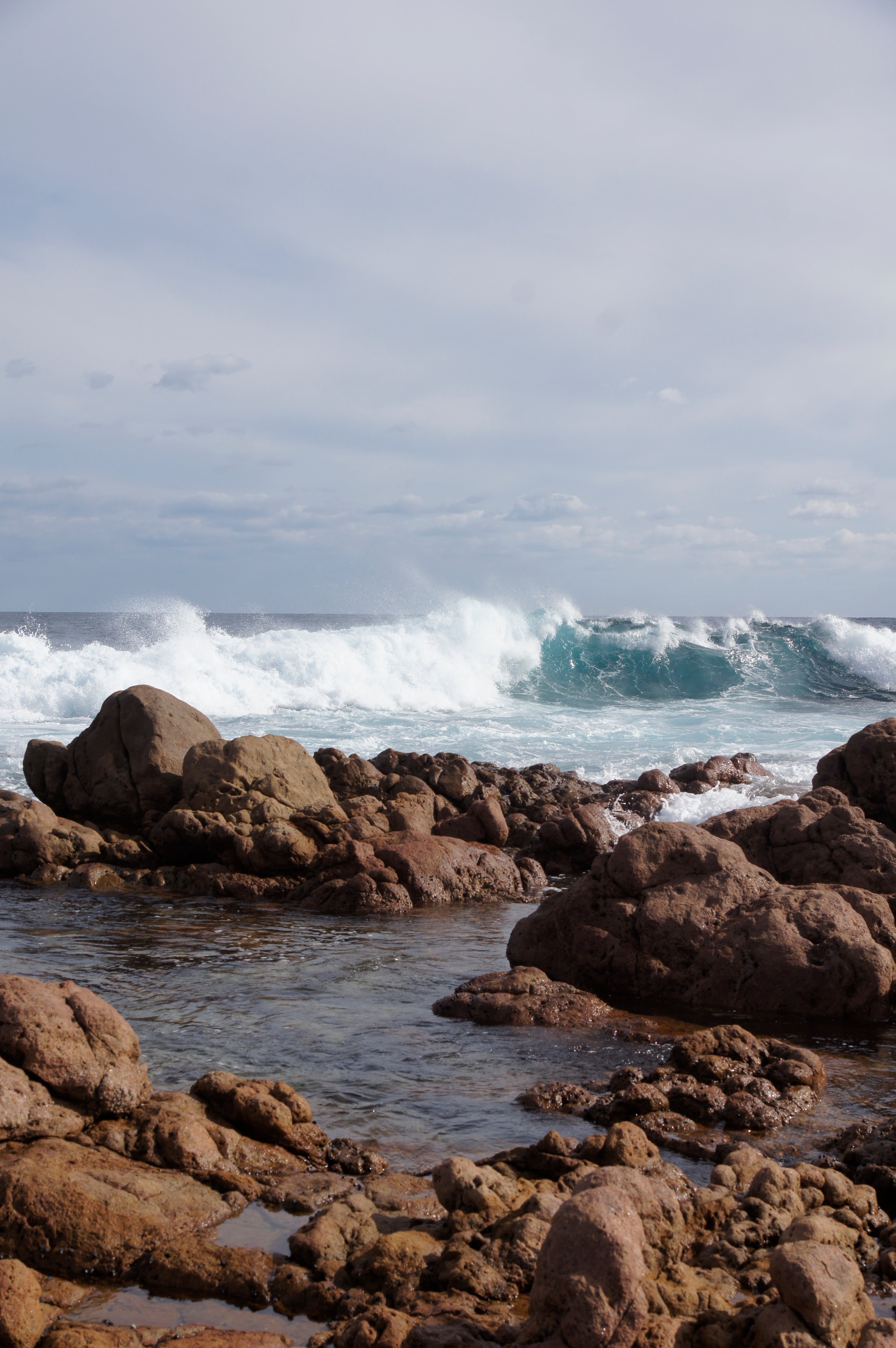 A rocky coastline with large brown rocks in the foreground and ocean waves breaking under a cloudy sky.