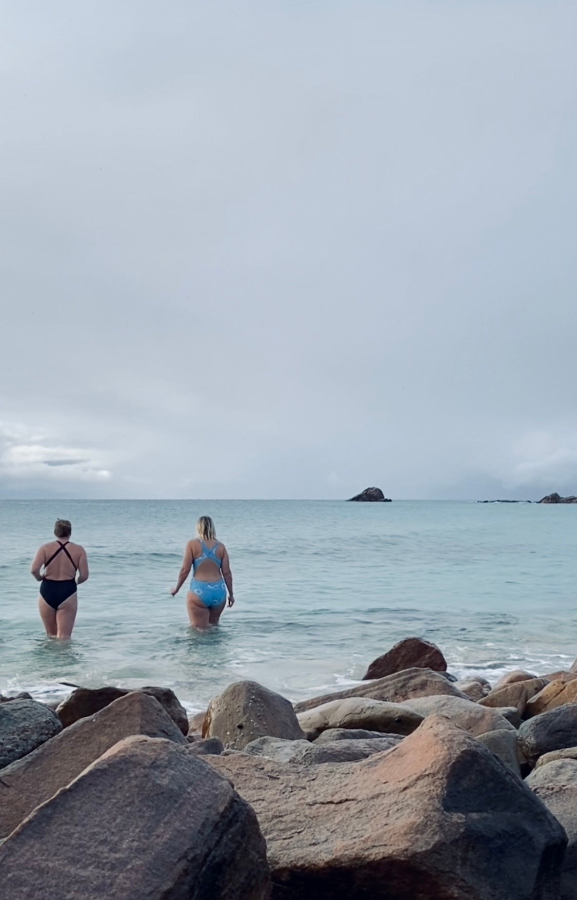 Two women in swimwear standing in shallow ocean water near a rocky shoreline under an overcast sky.