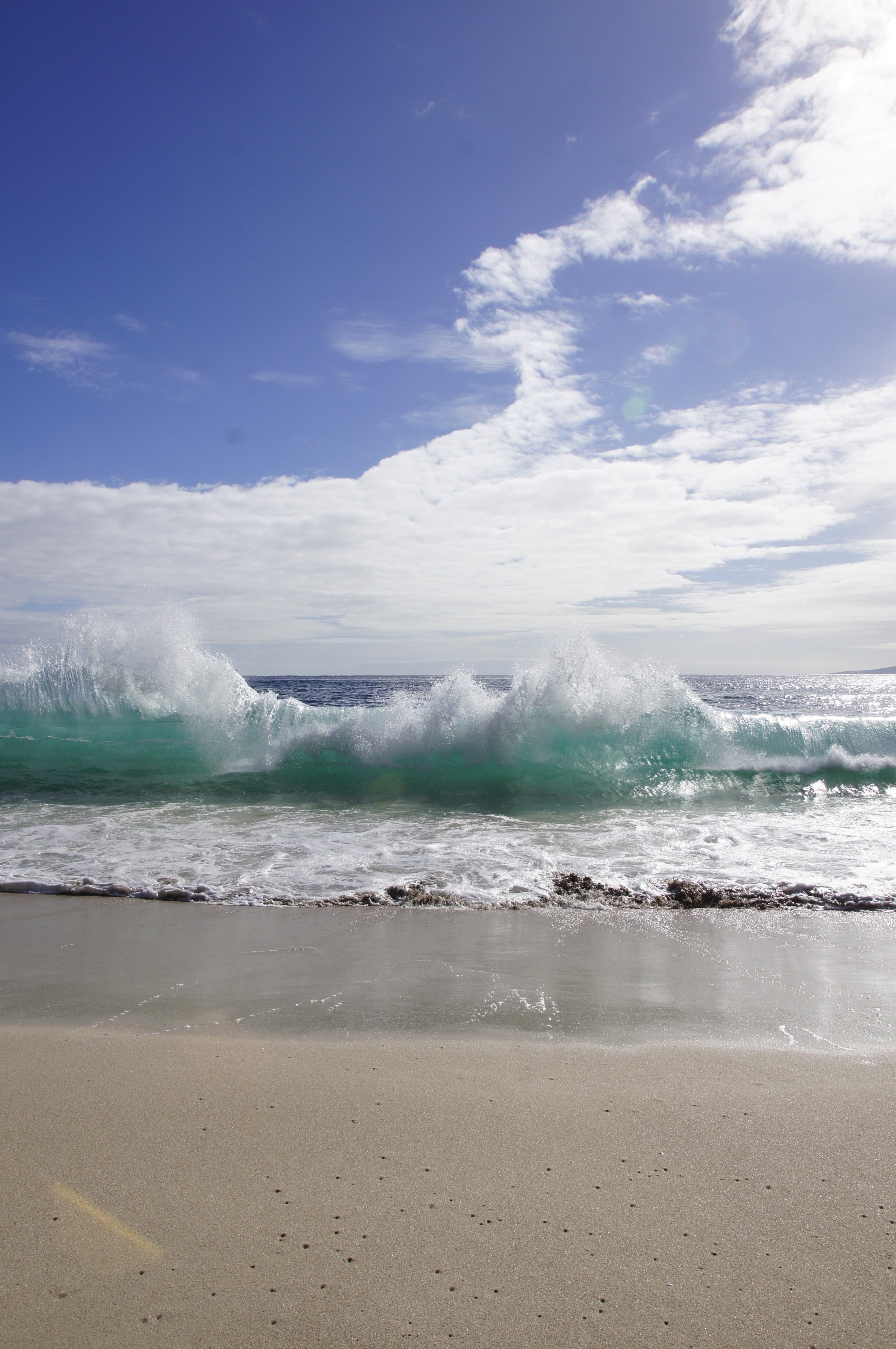 Beach scene with waves crashing onto the shore under a blue sky with clouds.