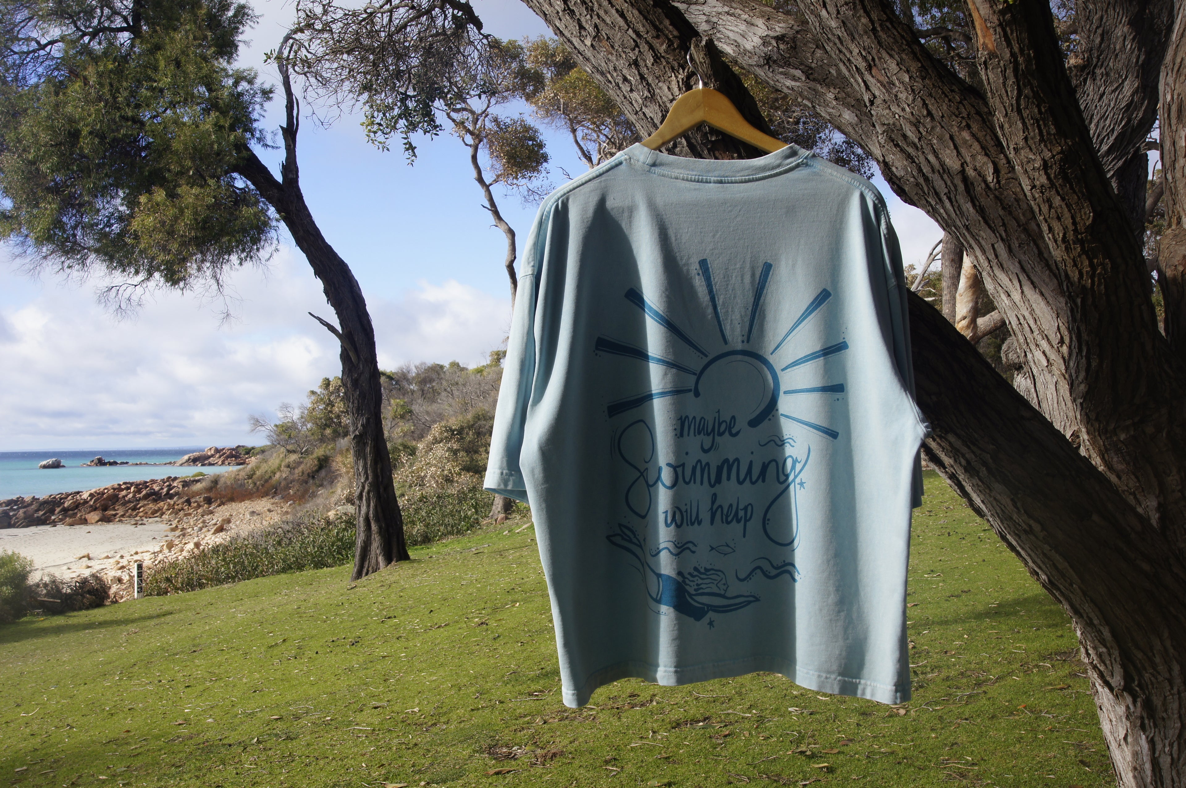 Light blue t-shirt with a sun design and text hanging on a tree branch by a beach.