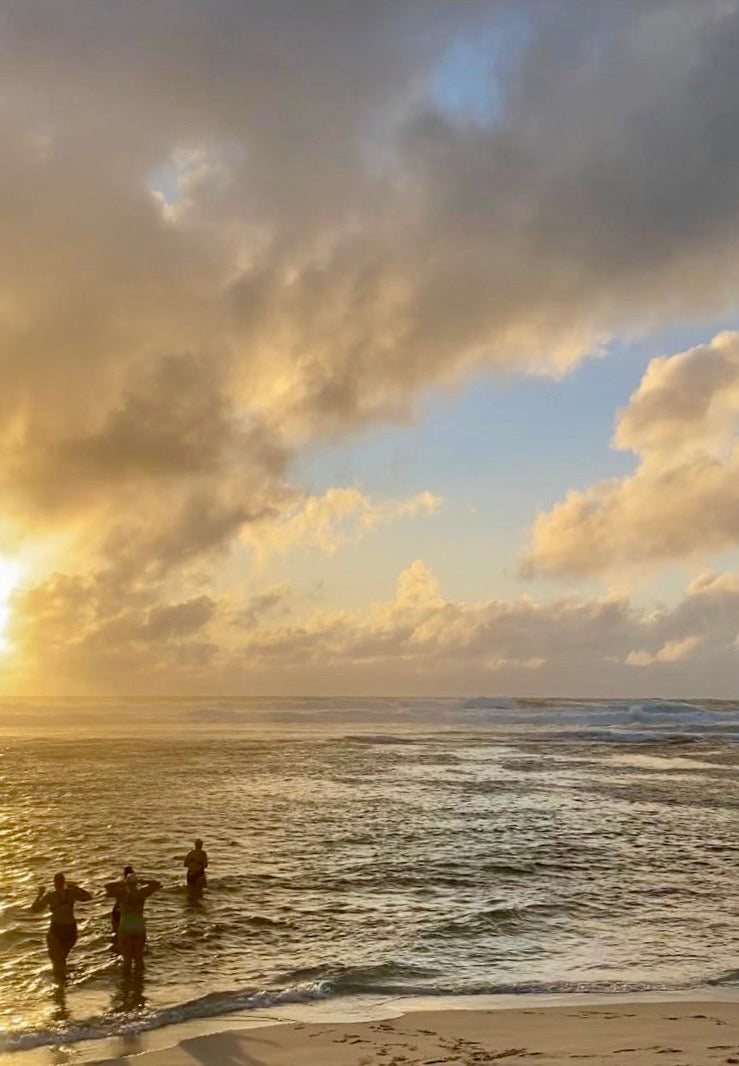 A beach sunset scene with ocean waves, dramatic clouds, and three silhouetted people standing in shallow water near the shore.